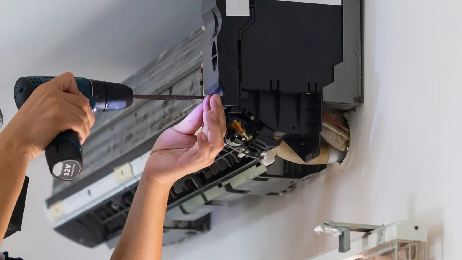 Hands using a cordless drill on the opened casing of an indoor air conditioning unit